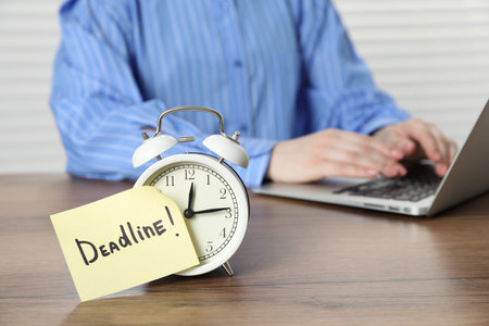 Deadline. Person with laptop working at wooden table in office, focus on alarm clock and sticky noteの写真素材