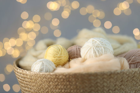 Soft balls of yarn and felting wool in wicker basket against gray background with blurred lights,...
