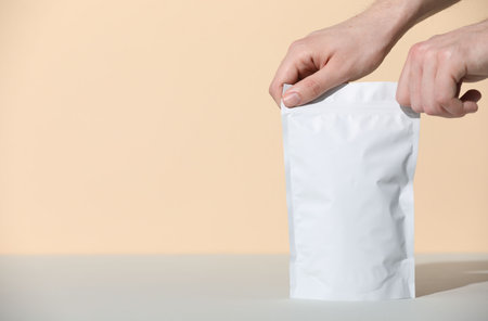 Woman holding blank zipper stand-up pouch at gray table against beige background, closeup. Space for...
