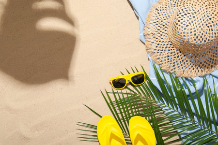 Beach towel, straw hat, sunglasses, flip flops and palm leaves on sand outdoors, flat lay. Space for...