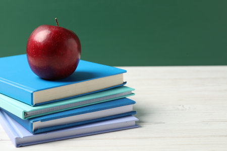 Different notebooks and red apple on white wooden table against blackboard, closeup. Space for textの写真素材
