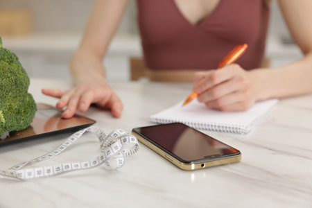 Woman developing diet plan while weighing broccoli on kitchen scale at white marble table indoors, closeupの写真素材
