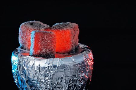 Smoldering charcoal cubes on hookah holder covered with foil against black background, closeup