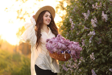 Smiling woman with basket of lilac flowers near bush outdoors in morningの写真素材