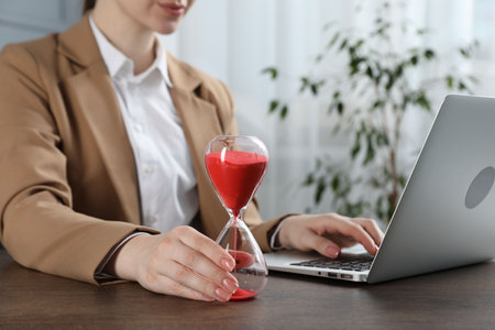 Deadline. Woman with hourglass and laptop working at wooden table in office, closeupの写真素材