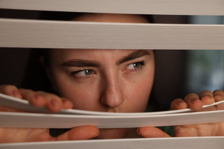 Paranoia. Worried woman looking through window blinds indoors, closeup