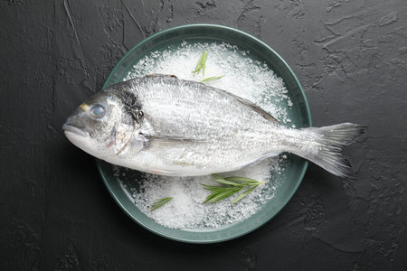 Fresh raw dorado fish, salt and rosemary in bowl on black table, top view. Organic seafood
