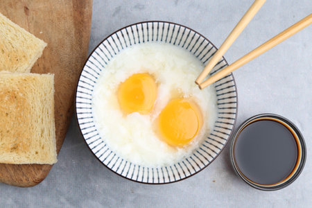 Traditional asian breakfast with half-boiled eggs in bowl, soy sauce, bread and chopsticks on grey...
