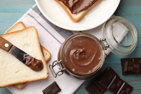 Tasty chocolate butter in jar, pieces, knife and bread on light blue wooden table, flat layの写真素材