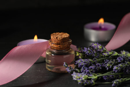 Bottle of lavender essential oil, flowers, burning candles and ribbon on black table, closeup