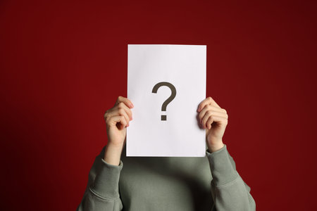 Anonymity. Woman hiding behind paper sheet with question mark on dark red backgroundの写真素材