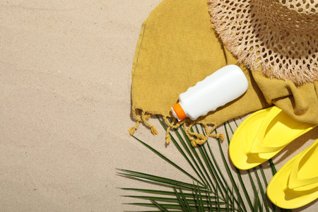 Beach towel, straw hat, flip flops, sunscreen product and palm leaf on sand outdoors, flat lay....