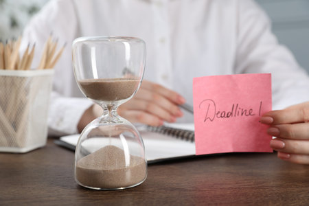 Woman holding sticky note with word Deadline at wooden table in office, closeupの写真素材