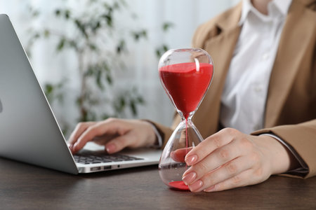 Deadline. Woman with hourglass and laptop working at wooden table in office, closeupの写真素材