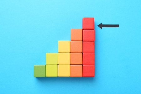 Maximum. Colorful cubes and arrow pointing at red one on light blue background, flat lay