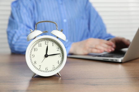 Deadline. Person with laptop working at wooden table in office, focus on alarm clock
