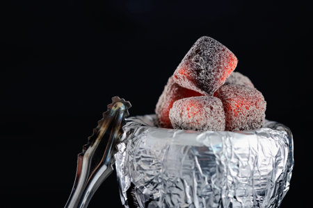 Smoldering charcoal cubes on hookah holder covered with foil and tongs against dark background, closeupの写真素材
