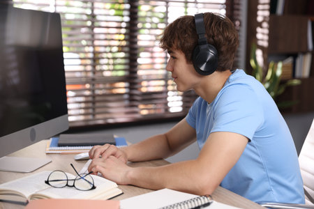 Teenage student with headphones using computer while studying at table indoors