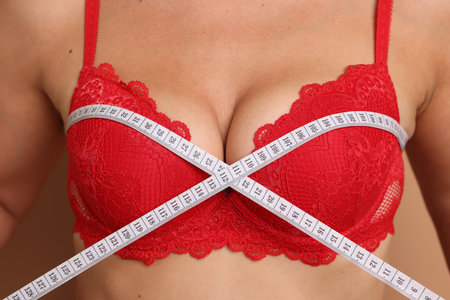 Woman in beautiful bra measuring breast circumference on beige background, closeupのeditorial素材