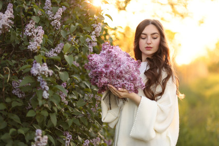 Beautiful woman with bouquet of lilac flowers near bush outdoors in morningの写真素材