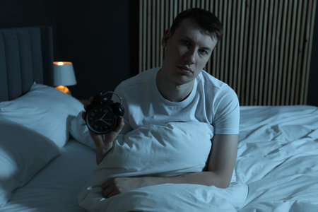 Frustrated man with alarm clock suffering from insomnia on bed indoorsの写真素材