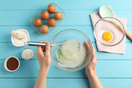 Woman whisking egg whites in bowl at light blue wooden table, top view