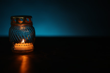 Grave lantern with burning candle on black table against blue background. Space for text