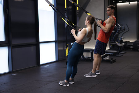 Young woman exercising with suspension trainers with professional personal coach in gym, space for...