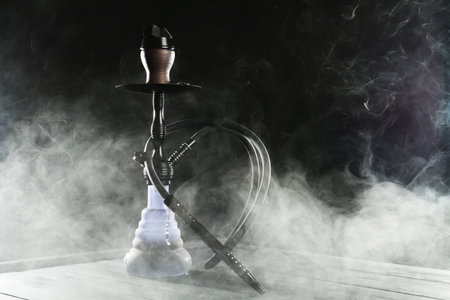 Hookah on wooden table with smoke against black background