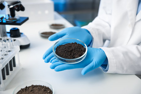 Scientist holding Petri dish with soil sample at white table indoors, closeup