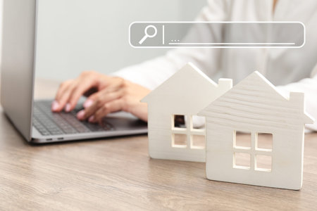 Searching for housing online. Real estate agent using laptop at table, closeup. Search bar above house modelsの写真素材