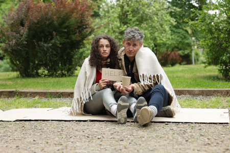 Homeless people with sign Help and paper cup on carton in parkの写真素材