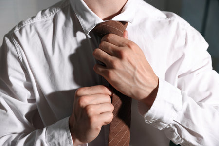 Man adjusting necktie while getting ready indoors, closeup