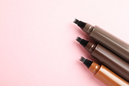 Liquid eyebrow pencils on light pink background, flat lay. Space for text