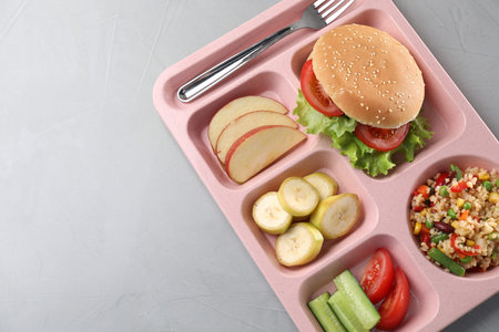 School lunch. Plastic tray with tasty food and fork on gray table, top view. Space for text