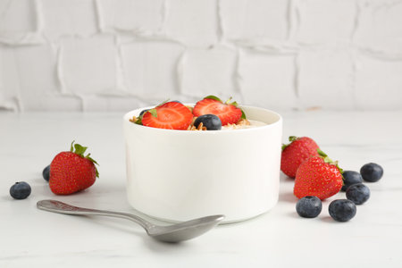 Tasty yogurt with granola, berries and spoon on white marble table, closeup