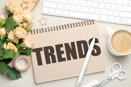 Trends inscription in notebook, flowers, computer keyboard, coffee and stationery on beige table,...
