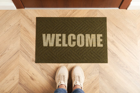 Woman standing in front of Welcome floor mat near door, top view