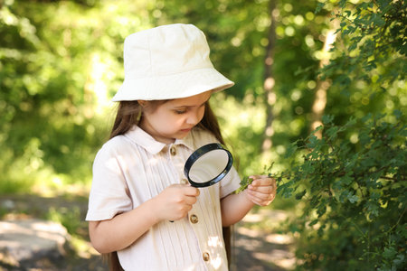 Cute little girl looking at branch with green leaves through magnifying glass in natureの写真素材