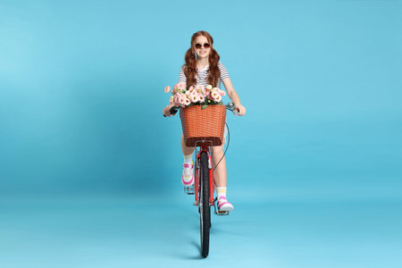 Smiling teenage girl in sunglasses riding bicycle with basket of flowers against light blue...