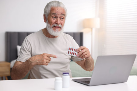 Senior man with pills having online consultation with doctor via laptop at table indoorsの写真素材