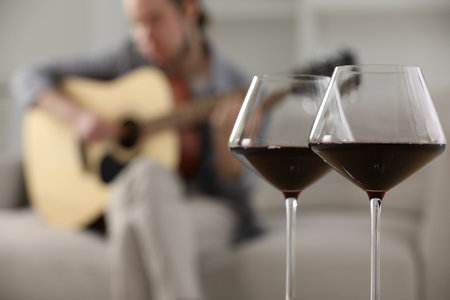 Glasses with red wine and man playing guitar indoors, selective focusの写真素材