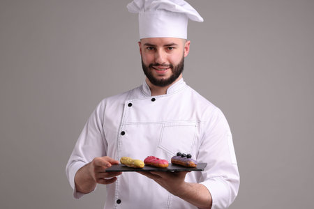 Happy confectioner in uniform holding delicious eclairs on light grey backgroundの写真素材