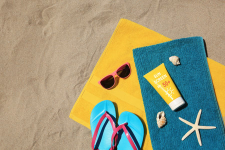 Sunscreens, flip flops, sunglasses and towels on sand, top view. Sun protection product