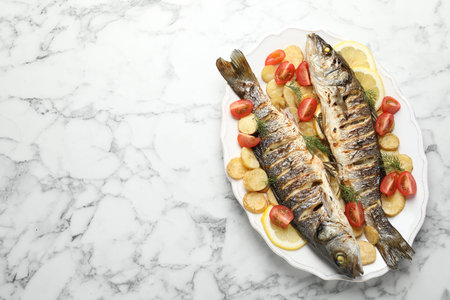 Tasty roasted seabass fish served with potatoes, tomatoes and lemon on white marble table, top view...