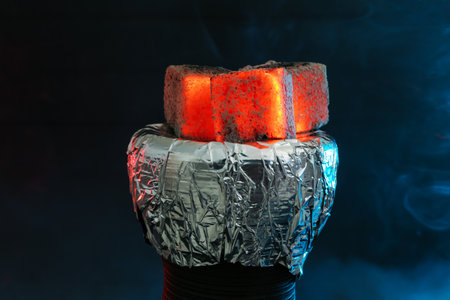 Smoldering charcoal cubes on hookah holder covered with foil and smoke in color light against dark background, closeupの写真素材