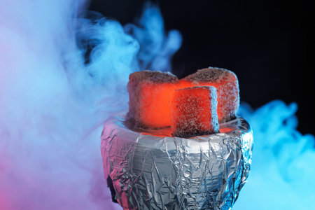 Smoldering charcoal cubes on hookah holder covered with foil and smoke in color lights against dark background, closeupの写真素材