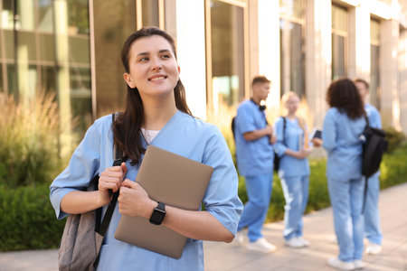 Happy medical student in uniform with backpack and laptop near building outdoors, selective focusの写真素材