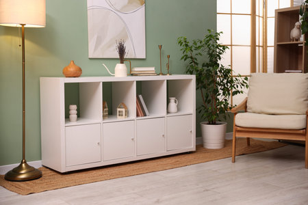 Shelving unit with decorative elements, lamp, houseplant and armchair in stylish room. Interior...