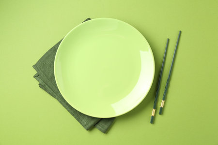 Stylish plate, chopsticks and napkin on green background, flat lay